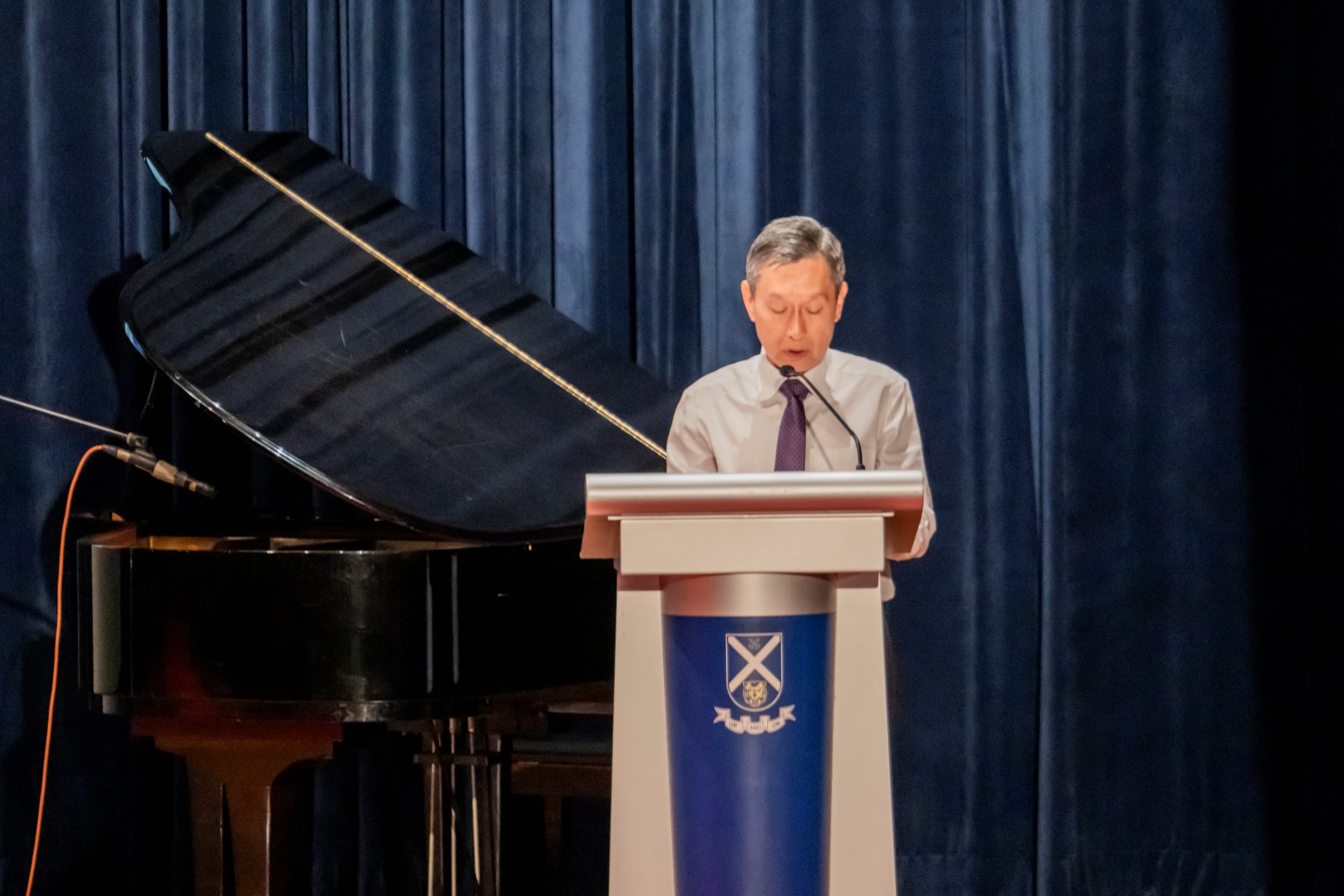 Deputy Principal, Mr Cecil Ang, in a white dress shirt and dark tie speaking at a podium beside an open grand piano on stage.